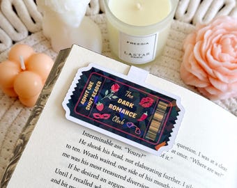 Könnte beinhalten: Ein Aufkleber mit dunkelblauem Hintergrund und einem rot-weiß-schwarzen Ticketdesign. Der Aufkleber lautet "The Dark Romance Club" und zeigt eine Erdbeere, eine Chilischote und ein Herz. Der Aufkleber befindet sich auf einem Buch mit Text über einen Charakter namens Wrath.