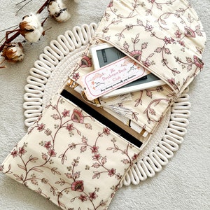 May include: A cream-colored fabric book sleeve with a floral pattern. The sleeve is open and a white e-reader is inside. A pink bookmark with the text "The Daydream Book Club" is sticking out of the e-reader. The text on the bookmark reads "Enjoy sharing your monthly grey characters."