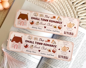 May include: A rectangular paper bookmark with a pale pink background featuring a ticket-style design. It displays the text "The Small Town Romance Book Club," illustrations of a house, picnic baskets, and a teapot, with a tassel attached to the top.