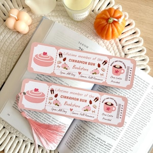 Könnte beinhalten: Zwei rechteckige Lesezeichen aus Papier im rosa-weißen Ticket-Design mit dem Text "Lifetime member of the Cinnamon Bun Bookstore". Die Lesezeichen zeigen Illustrationen einer Zimtschnecke, eines Chai Latte und Kürbissen sowie eine rosa Quaste.