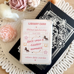 May include: A clear, rectangular library card with the text "LIBRARY CARD" and "ONCE UPON A COWBOY ROMANCE." The card is in a clear case, resting on a black book with floral cutouts. Decorative candles and flowers surround the card.