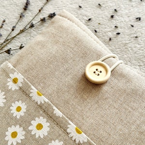 May include: A beige linen pouch with a white daisy print and a wooden button closure. The pouch is on a white fuzzy surface.
