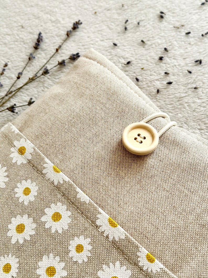 May include: A beige linen pouch with a white daisy print and a wooden button closure. The pouch is on a white fuzzy surface.