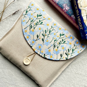 Può includere: Una pochette in tela beige con chiusura a lembo e bottone. Il lembo è decorato con fiori ricamati blu e gialli. La pochette è appoggiata su una superficie bianca.