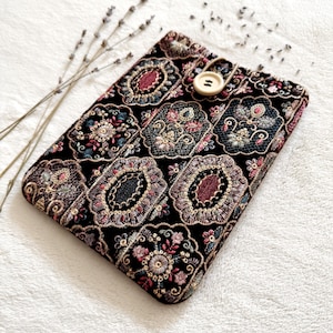 May include: Black tablet case with an ornate floral pattern in shades of pink, red, green, and gold. The case has a button closure and is decorated with a detailed embroidered design. A sprig of lavender is placed near the case.