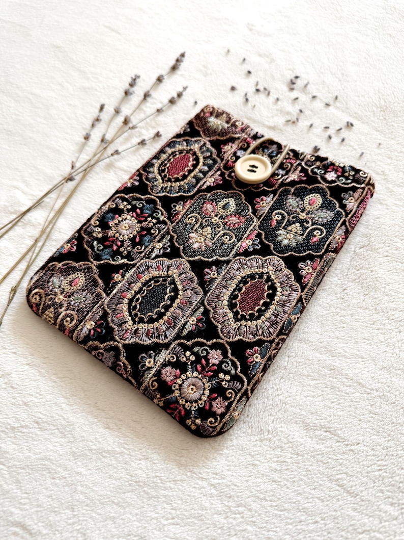 May include: Black tablet case with an ornate floral pattern in shades of pink, red, green, and gold. The case has a button closure and is decorated with a detailed embroidered design. A sprig of lavender is placed near the case.