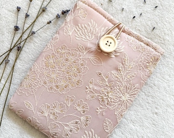 Puede incluir: Una funda para tablet de tela rosa con un patrón floral blanco y un cierre de botón blanco.