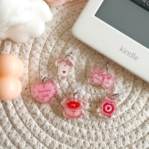 Op de afbeelding: Vijf roze en doorzichtige plastic boekenleggers met verschillende ontwerpen. De ontwerpen omvatten een hart met de tekst "Smells Like Smut", een hart met de tekst "Romantary Girlie", een taart met de tekst "I wish for more books", een bril met de tekst "Bookish Babe" en een pot met de tekst "Reading".