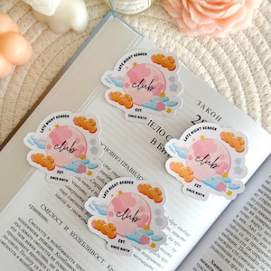Könnte beinhalten: Vier Aufkleber mit einem rosa Mond-Design, dem Wort "club" und dem Text "Late Night Reader" und "Since Birth". Die Aufkleber befinden sich auf einem offenen Buch mit Text, umgeben von Kerzen und einer dekorativen Blume.