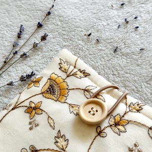 May include: A white fabric pouch with a floral embroidery design in gold and brown. The pouch has a wooden button closure and a brown elastic strap.