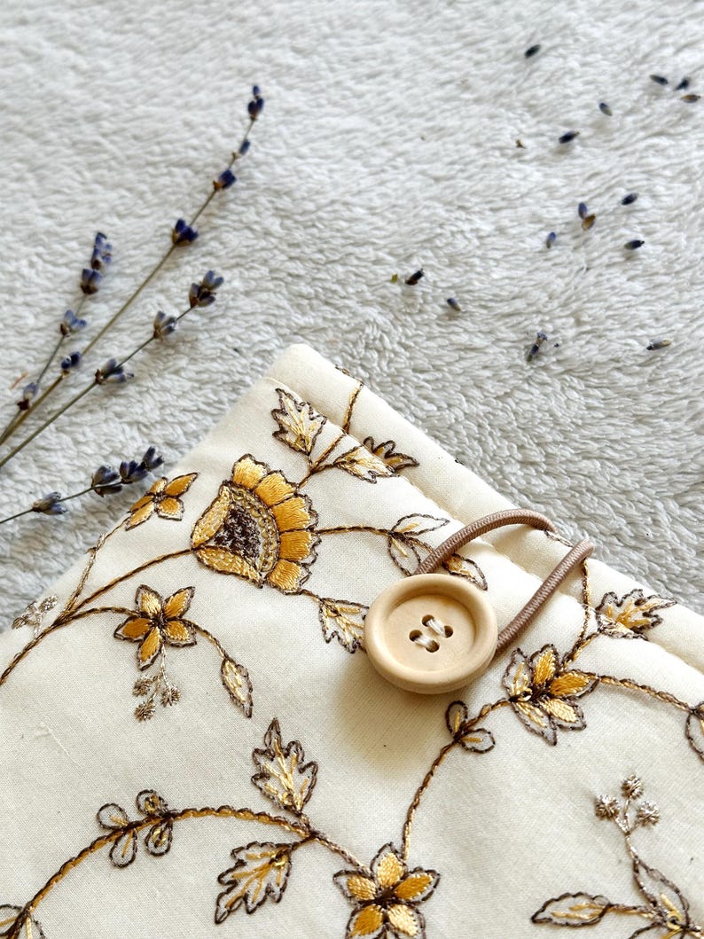 May include: A white fabric pouch with a floral embroidery design in gold and brown. The pouch has a wooden button closure and a brown elastic strap.