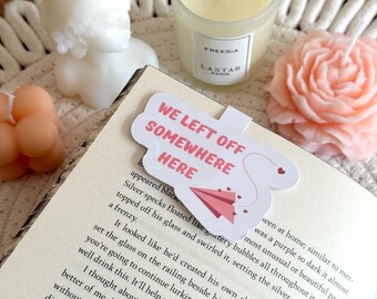 May include: A still-life arrangement featuring a variety of candles and a bookmark. The candles include a white bust, a clear glass candle with a label, a peach-colored rose, and a small peach-colored cluster. The bookmark reads "WE LEFT OFF SOMEWHERE HERE".