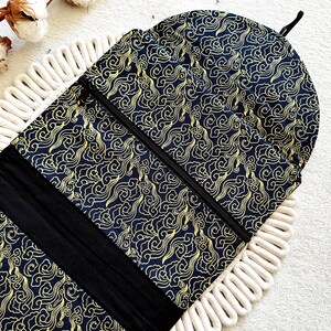 May include: A black prayer rug with a gold cloud pattern and a black zipper. The rug is rolled up and has a black strap with a hook.