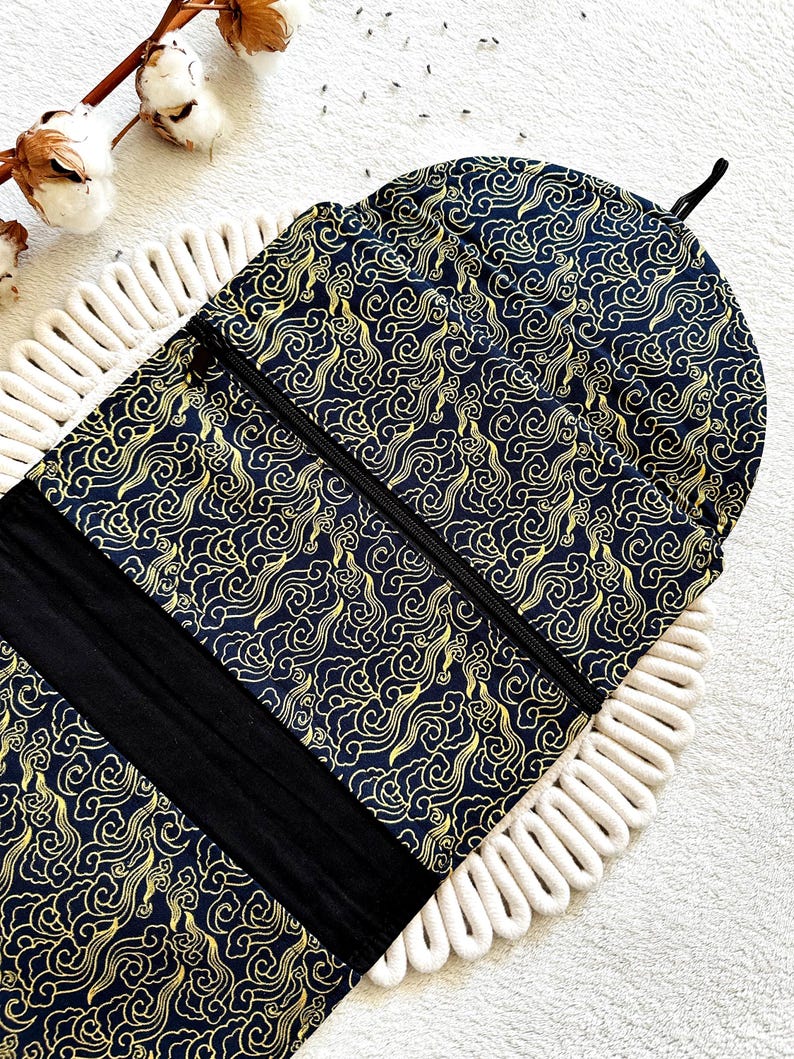 May include: A black prayer rug with a gold cloud pattern and a black zipper. The rug is rolled up and has a black strap with a hook.