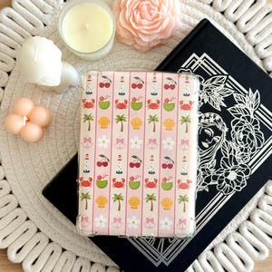 May include: A pink and white patterned tablet case with a summer theme, featuring cherries, crabs, coconuts, palm trees, and seashells. The case is on a black book, surrounded by candles and a decorative mat.