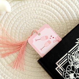 May include: A rectangular pink bookmark featuring a graphic of two champagne flutes and small red hearts, finished with a light pink tassel. The bookmark is inserted into a black book with a white floral design on the cover.