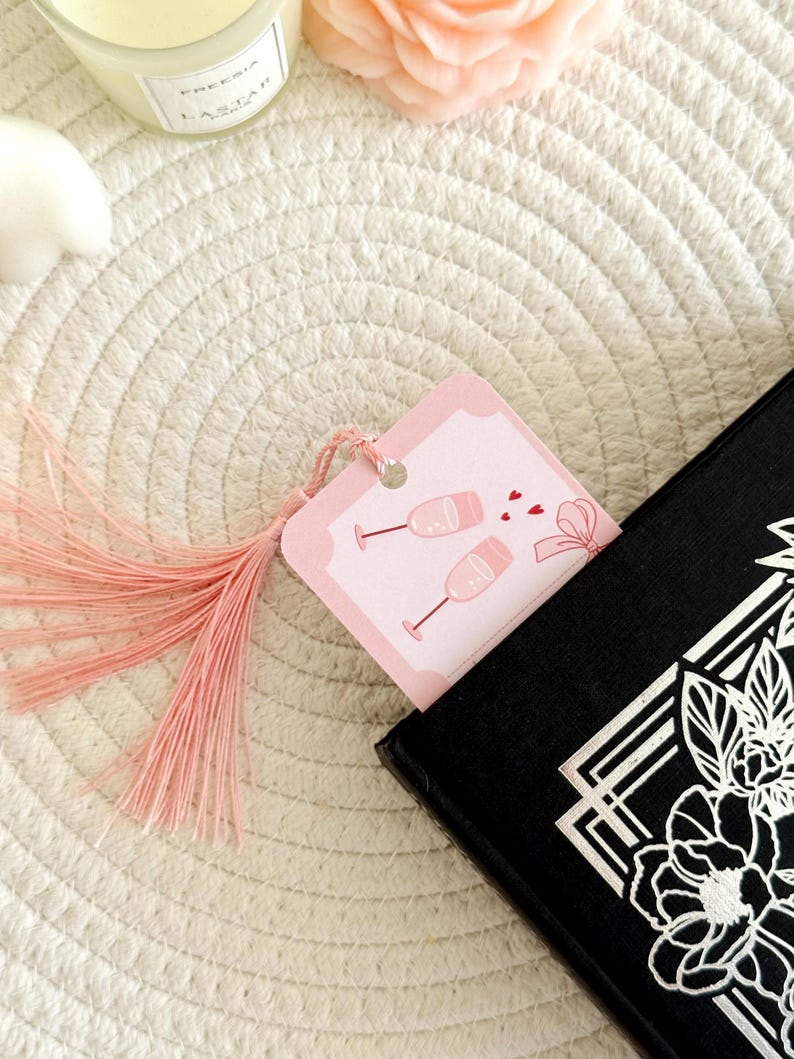 May include: A rectangular pink bookmark featuring a graphic of two champagne flutes and small red hearts, finished with a light pink tassel. The bookmark is inserted into a black book with a white floral design on the cover.
