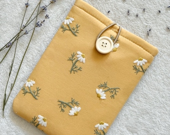 Puede incluir: Una funda de teléfono de tela amarilla con cierre de botón blanco y un patrón floral de margaritas blancas bordadas.