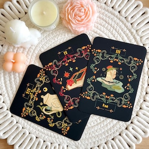 May include: Three black tarot cards with gold floral designs and illustrations are arranged on a white woven mat. The cards feature images of a book, a figure in water, and a creature. Candles and a rose-shaped candle are also present.
