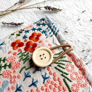 May include: A small, beige fabric pouch with a floral embroidery design. The pouch has a brown elastic strap and a wooden button closure. The embroidery features pink, orange, green, and blue flowers and leaves.