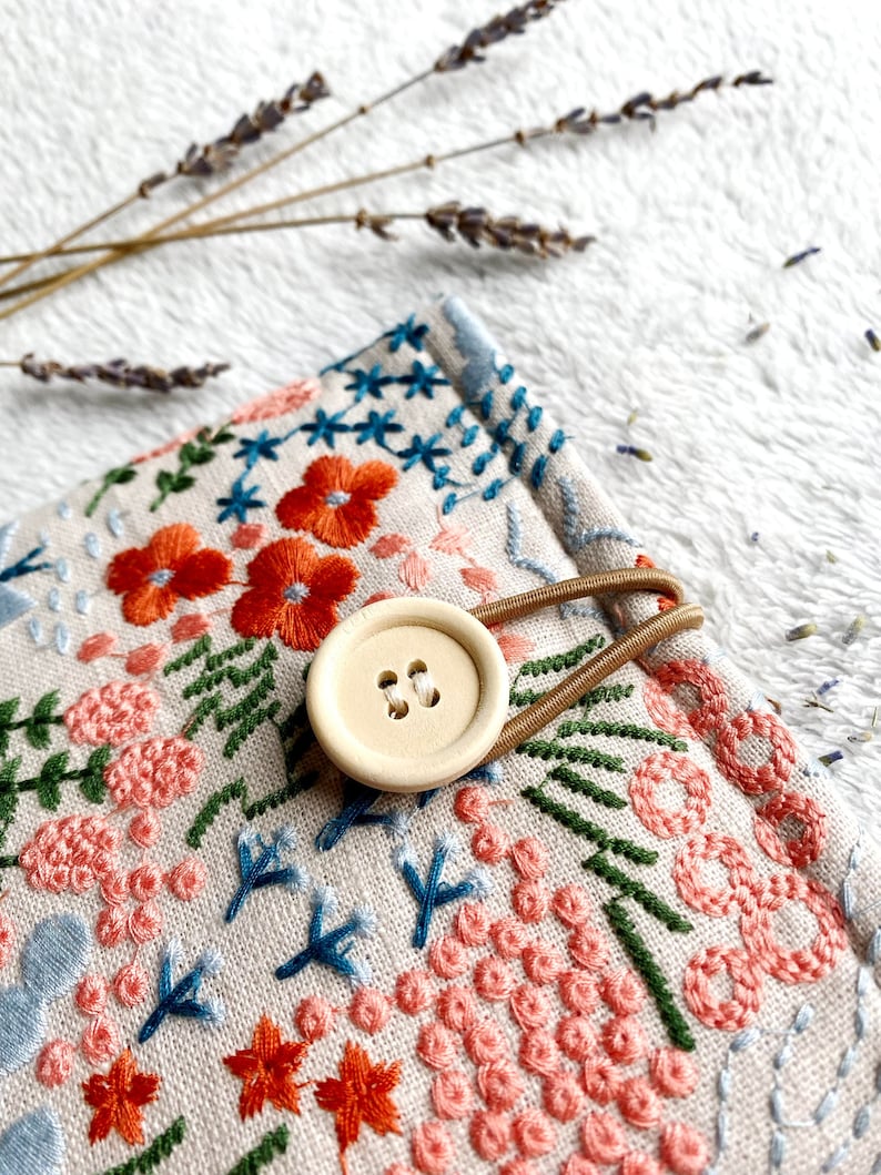 May include: A small, beige fabric pouch with a floral embroidery design. The pouch has a brown elastic strap and a wooden button closure. The embroidery features pink, orange, green, and blue flowers and leaves.