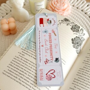 May include: A bookmark with a light blue tassel rests on an open book. The bookmark features illustrations of books, a mug, and a basketball, with the text "The Forced Proximity Book Club."