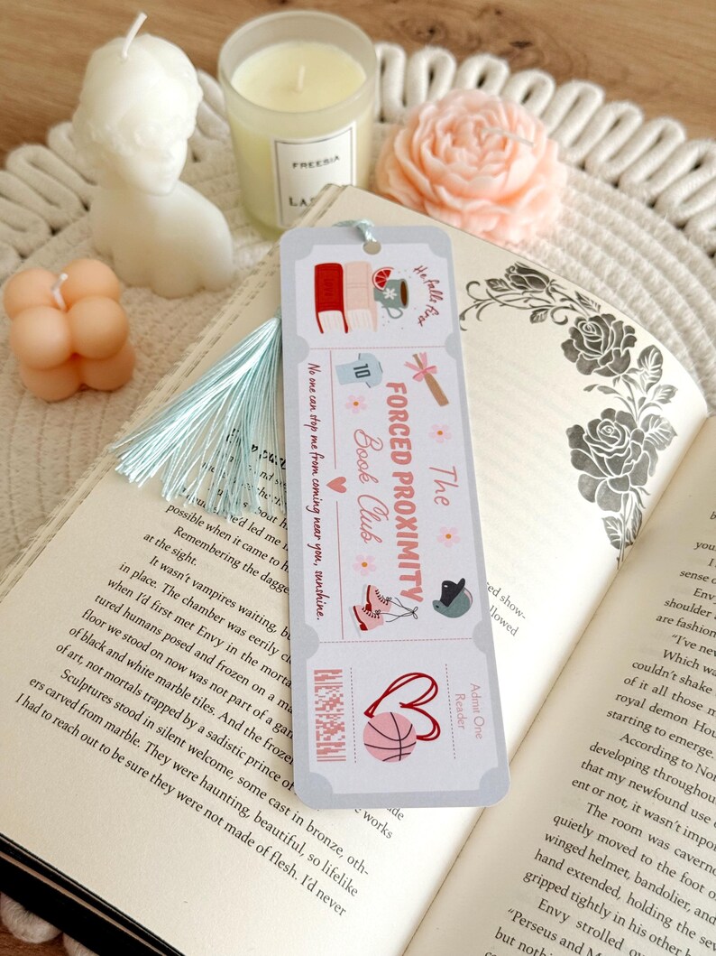 May include: A bookmark with a light blue tassel rests on an open book. The bookmark features illustrations of books, a mug, and a basketball, with the text "The Forced Proximity Book Club."