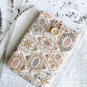 Könnte beinhalten: Eine rechteckige, gemusterte Buchhülle mit Knopfverschluss. Die Hülle zeigt ein sich wiederholendes Design aus floralen Medaillons in Creme, Gold, Rosa und Blau. Lavendelzweige und -knospen befinden sich im Hintergrund.