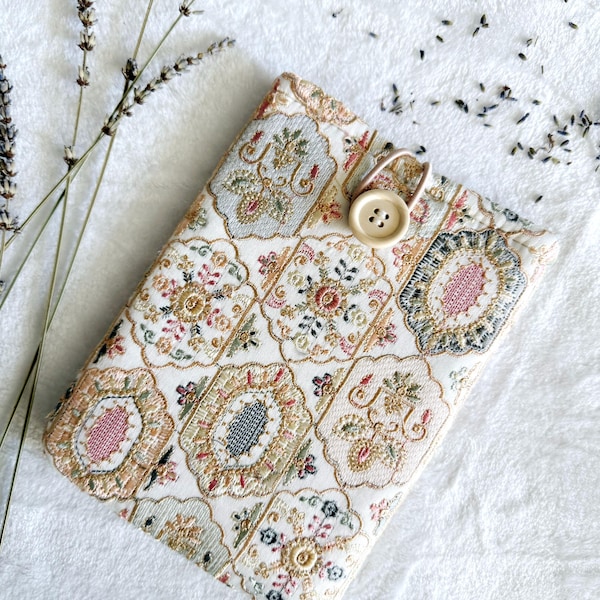 Könnte beinhalten: Eine rechteckige, gemusterte Buchhülle mit Knopfverschluss. Die Hülle zeigt ein sich wiederholendes Design aus floralen Medaillons in Creme, Gold, Rosa und Blau. Lavendelzweige und -knospen befinden sich im Hintergrund.