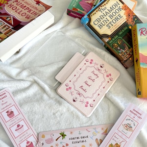 May include: A collection of books and bookmarks on a white surface. The books include titles like "Love and Lattes at Pumpkin Hollow" and "The Cinnamon Bun Book Store." Bookmarks feature phrases like "Coastal Girlie Essentials" and "Summer Bucket List."