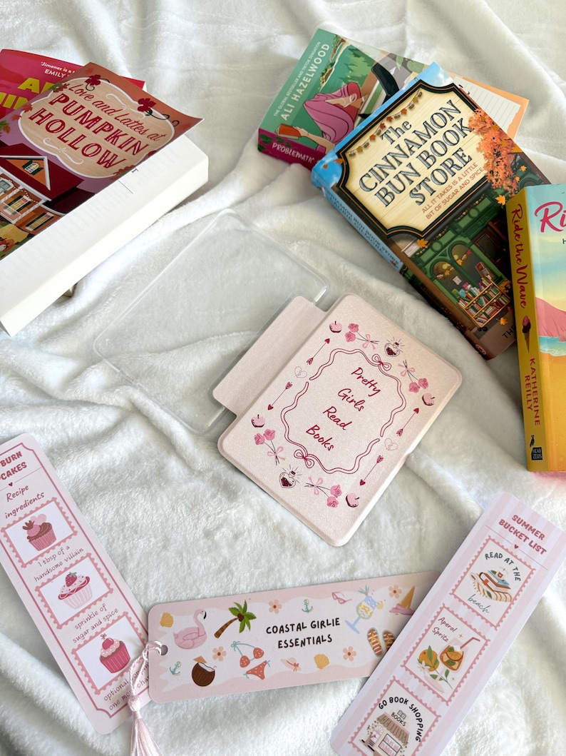 May include: A collection of books and bookmarks on a white surface. The books include titles like "Love and Lattes at Pumpkin Hollow" and "The Cinnamon Bun Book Store." Bookmarks feature phrases like "Coastal Girlie Essentials" and "Summer Bucket List."