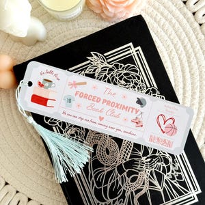May include: A bookmark with a light blue tassel rests on a black book with a silver floral design. The bookmark reads "The Forced Proximity Book Club" with illustrations of books, a cup, and a basketball.
