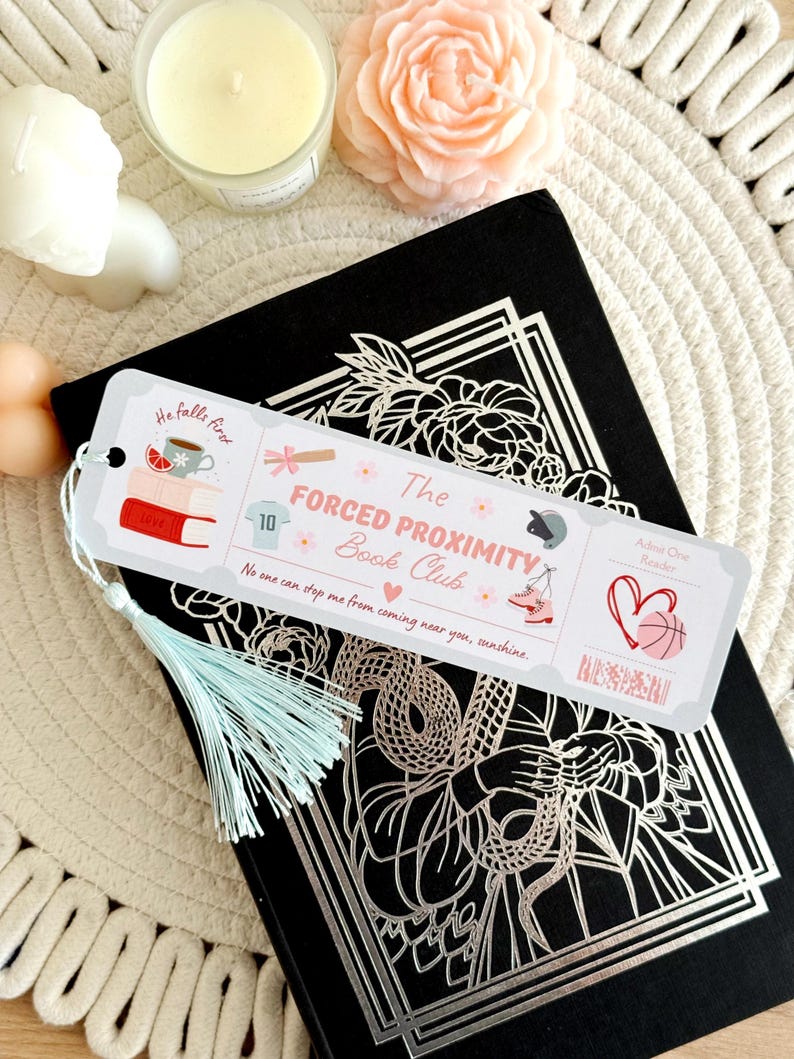 May include: A bookmark with a light blue tassel rests on a black book with a silver floral design. The bookmark reads "The Forced Proximity Book Club" with illustrations of books, a cup, and a basketball.