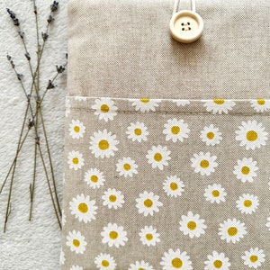 May include: A beige linen pouch with a white daisy print and a single button closure. The pouch is made of natural fabric and has a simple, elegant design.