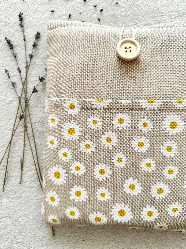 May include: A beige linen pouch with a white daisy print and a single button closure. The pouch is made of natural fabric and has a simple, elegant design.
