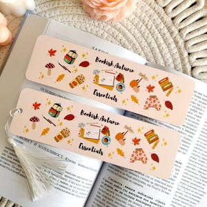 Puede incluir: Dos marcapáginas rectangulares de papel con fondo color melocotón claro, con ilustraciones coloridas de elementos otoñales como calabazas, hojas, libros y jerséis, con el texto Bookish Autumn Essentials en letra cursiva negra.