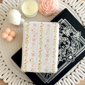 Peut inclure: Une coque de téléphone transparente avec un motif d'oies blanches, de nœuds roses et de tiges florales vertes. La coque est posée sur un livre noir avec un motif floral et un dessous de plat blanc tressé. Des bougies et une fleur décorative sont également visibles.