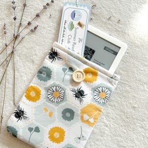 May include: A white and blue fabric e-reader case with a bee and flower pattern. The case has a button closure and is decorated with a yellow and green honeycomb design. The text on the bookmark reads "The Summer Read Club When all else fails, take a"
