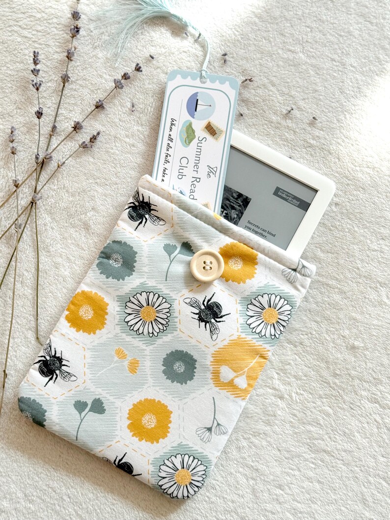 May include: A white and blue fabric e-reader case with a bee and flower pattern. The case has a button closure and is decorated with a yellow and green honeycomb design. The text on the bookmark reads "The Summer Read Club When all else fails, take a"