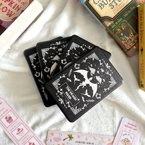 Könnte beinhalten: Vier schwarze Untersetzer mit einem weißen Drachen und dem Text "The Dragon River". Ebenfalls zu sehen sind Bücher und Lesezeichen, darunter eines mit der Aufschrift "Coastal Girlie Essentials".