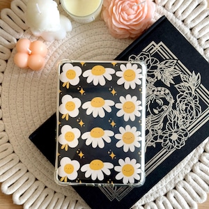 May include: A clear phone case with a black background and a pattern of white daisies with yellow centres and smiling faces. The daisies are scattered randomly across the case.