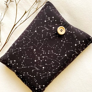 May include: A black fabric pouch with a white constellation pattern and a wooden button closure. The pouch is lying on a white surface.