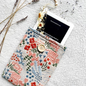May include: A beige fabric pouch with a floral embroidery design. The pouch has a button closure and is holding a white frame with a black background. The text on the frame reads "Recommended for you" and "Secrets & Demons (Demon Hunter in Hiding Book 1)".