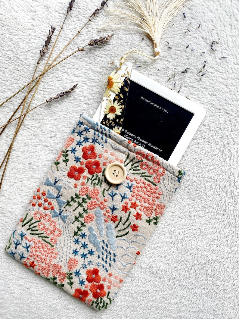 May include: A beige fabric pouch with a floral embroidery design. The pouch has a button closure and is holding a white frame with a black background. The text on the frame reads "Recommended for you" and "Secrets & Demons (Demon Hunter in Hiding Book 1)".