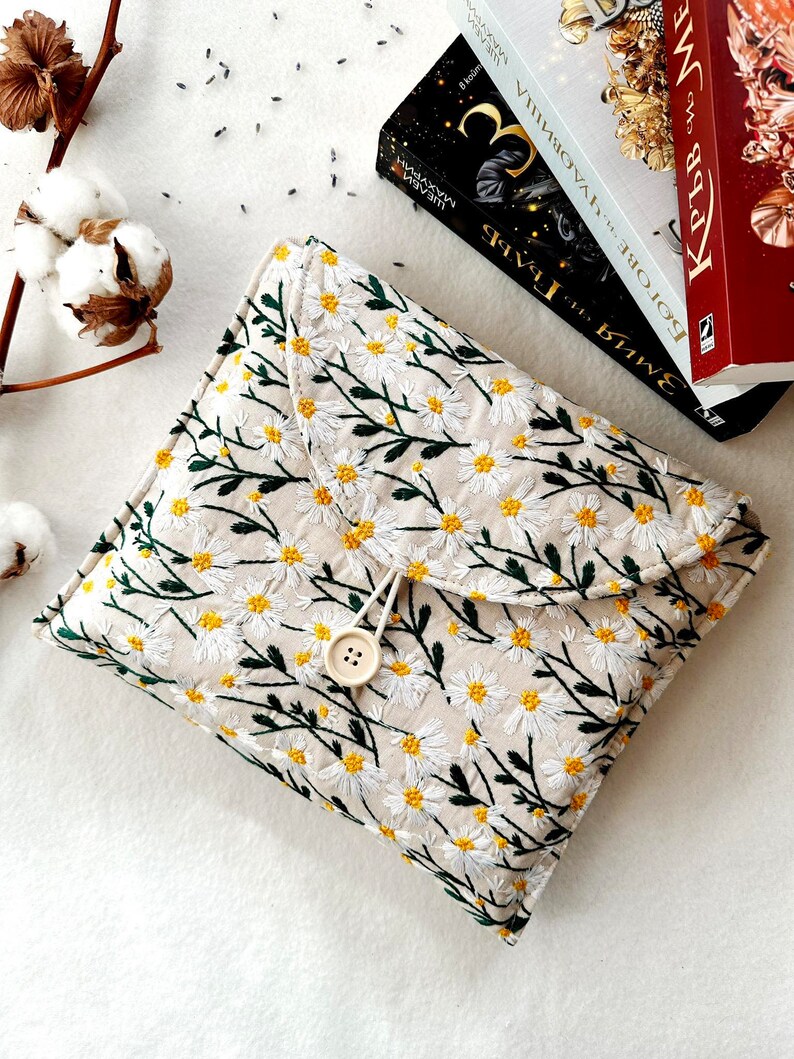 May include: A beige fabric clutch with a floral pattern of white daisies and green stems. The clutch has a flap closure with a button and a loop.