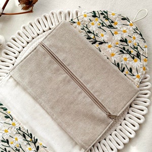 May include: A beige zippered pouch with a white cotton rope trim. The pouch is open to reveal a white fabric lining with a floral pattern of white daisies and green stems embroidered on it.