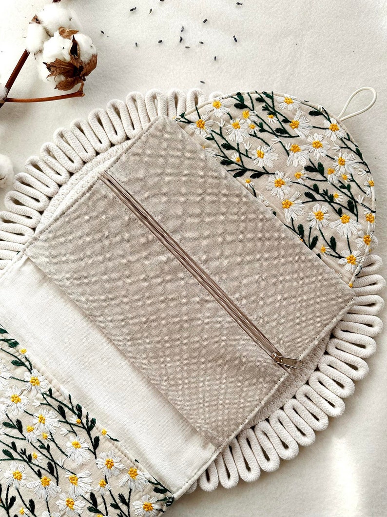 May include: A beige zippered pouch with a white cotton rope trim. The pouch is open to reveal a white fabric lining with a floral pattern of white daisies and green stems embroidered on it.
