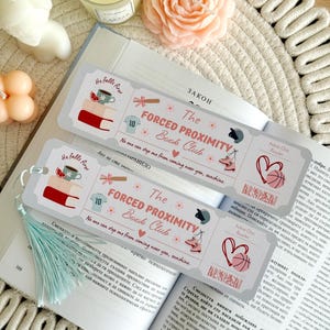 May include: Two decorative bookmarks with the text "The Forced Proximity Book Club" and illustrations of books, a cup, and sports equipment. The bookmarks are on an open book with a light blue tassel.
