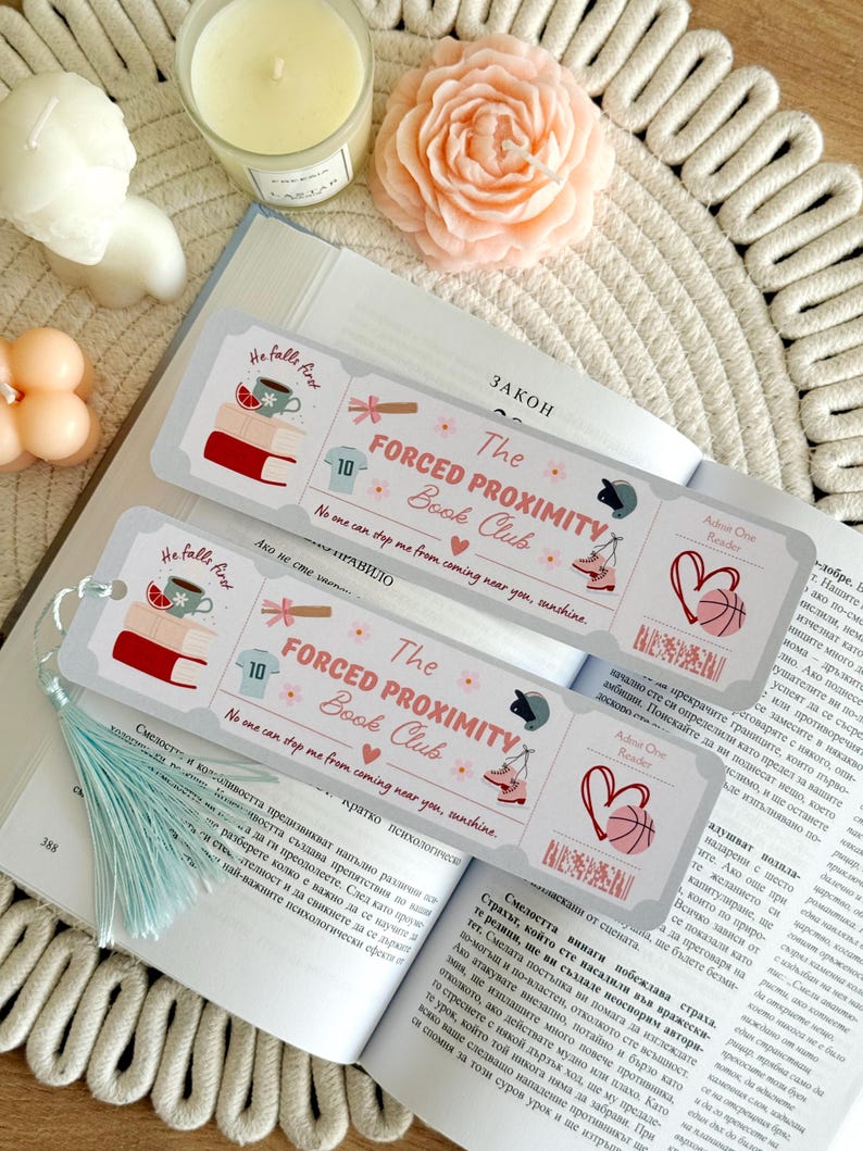 May include: Two decorative bookmarks with the text "The Forced Proximity Book Club" and illustrations of books, a cup, and sports equipment. The bookmarks are on an open book with a light blue tassel.