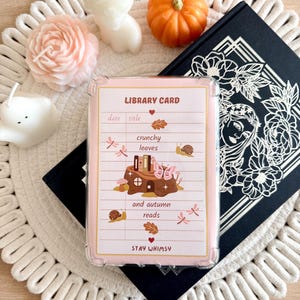 May include: A rectangular library card print in a clear protective sleeve featuring the text LIBRARY CARD, crunchy leaves, and autumn reads. The design includes illustrations of a tree stump, books, a butterfly, snails, and dragonflies.